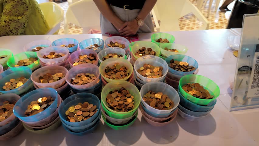 Continuous right-to-left traveling shot revealing dozens of colorful plastic bowls stacked with coins on a long donation table in a temple during a religious event