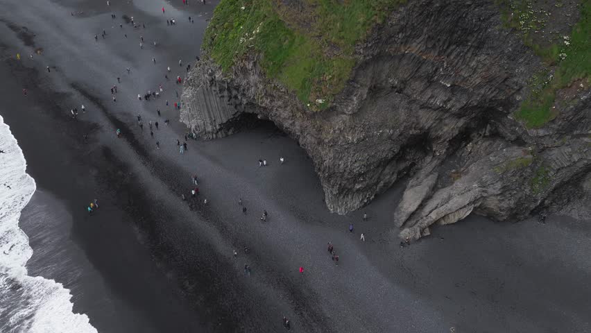 Aerial view of Reynisfjara Beach in Iceland, showing basalt columns, a large cave, visitors walking, and waves washing onto the black sand.