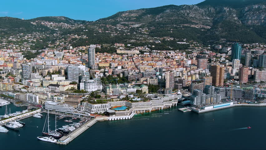 Glamorous drone panorama of Monaco’s Larvotto coast: luxury super‑yachts, Casino de Monte‑Carlo domes, high‑rise skyline and sheer Maritime Alps plunging into the sapphire Mediterranean Sea.