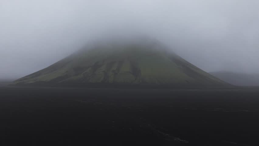 Aerial pan reveals a mist covered green mountain rising from a dark volcanic plain under overcast skies, showcasing Iceland