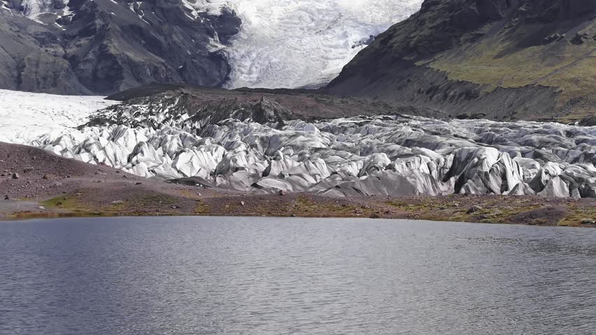 Aerial view of a glacier in Iceland, featuring dark volcanic ash streaks, rugged icy surfaces, and a mountainous backdrop with green patches.