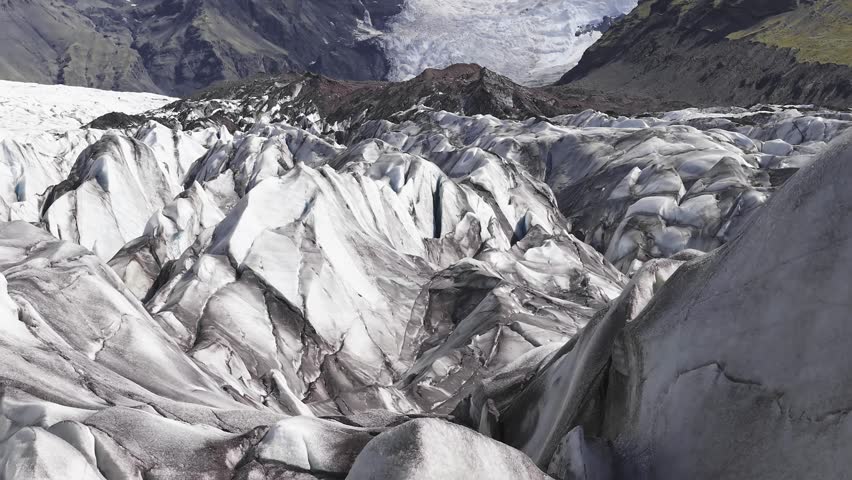 Aerial view of a glacier in Iceland, showcasing icy crevices, dark sediment streaks, rocky terrain, and distant mountains with slow camera motion.
