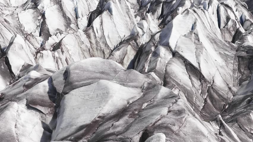 Sweeping aerial view of a glacier in Iceland, showcasing jagged ice formations, deep crevices, and steep snow covered mountains in the background.
