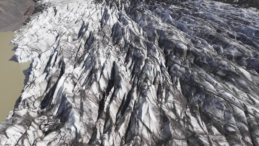A slow aerial sweep over a glacier in Iceland, showcasing icy crevasses, volcanic terrain, green cliffs, and rugged mountains under a cloudy sky.