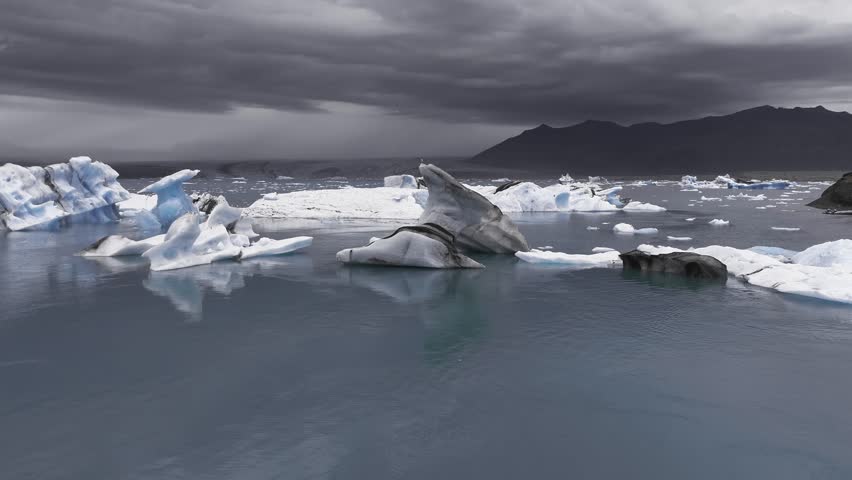Serene aerial view of Jokulsarlon Glacier Lagoon in Iceland, showing drifting icebergs, reflective water, dark mountains, and minimal human activity.