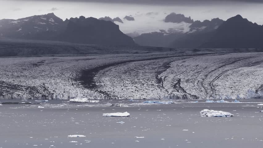 Aerial view of a Jokulsarlon glacier in Iceland with icebergs in a lagoon, jagged mountains in the background, and slow motion revealing the icy landscape.