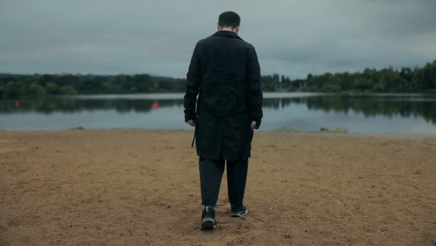 Young male wearing dark coat walks alone on sandy lake shore, overcast clouds above create a vibe of loneliness, sadness and inner reflection.