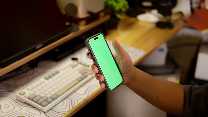 Hand holding a smartphone with green screen above a stylish keyboard and desk. A creative environment perfect for app demo, UI mockup, or digital workspace concept.