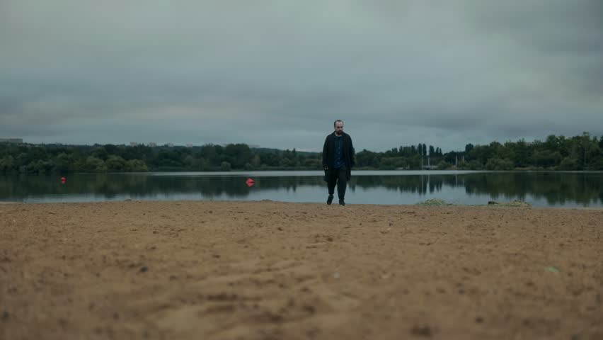 A middle-aged man walks slowly across a deserted beach in dark clothes, lost in thought. His slouched posture and somber expression convey deep sadness and isolation.