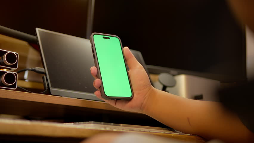 Close-up of a hand holding green screen smartphone in a modern tech workspace. Ideal for showcasing app interface, tech demo, or creative digital workflow presentation.