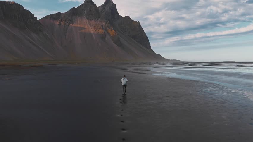 A person walks across the black sand beach at Stokksnes, Iceland, with Vestrahorn mountain in soft sunlight. Footprints and wet sand reflect the sky.