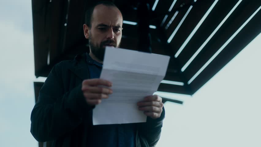 Bearded young man reads letter with visible sadness and confusion, standing outside in casual coat, overwhelmed by anxiety, shock and emotional pain.