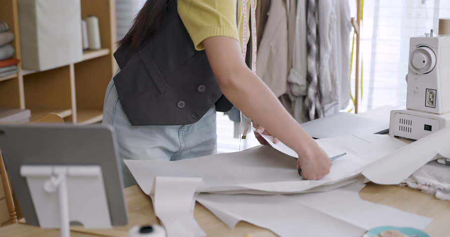 Asian young female fashion designer drawing cutting pattern lines on large white paper for handmade garment production in tailoring studio with measuring tape and tools placed on wooden desk