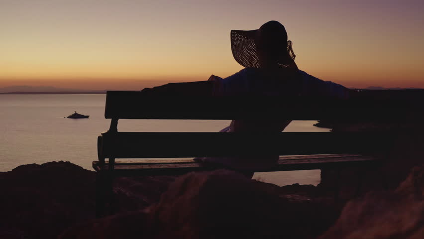 Cyprus: Silhouette of woman wearing summer hat relaxing on bench enjoying beautiful sunset view over Mediterranean Sea with distant boat in Cyprus. Drone flight footage
