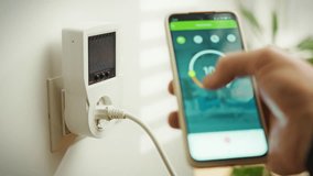 A person is interacting with a smart home energy monitoring device plugged into a wall outlet. The smartphone displays controls and data for managing energy consumption in real-time - Powered by Shutterstock - Get 15% off with code: PIKWIZARD15