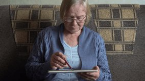 Senior woman wearing eyeglasses comfortably sitting on sofa at home. Female using digital pen while interacting with tablet, demonstrating digital literacy and modern lifestyle. Old peoples using apps - Powered by Shutterstock - Get 15% off with code: PIKWIZARD15