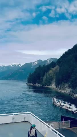 A breathtaking panoramic view of the Canadian wilderness featuring snow-covered mountains, dense evergreen forests, and a calm glacial lake. Captured under natural daylight with dramatic clouds, this 