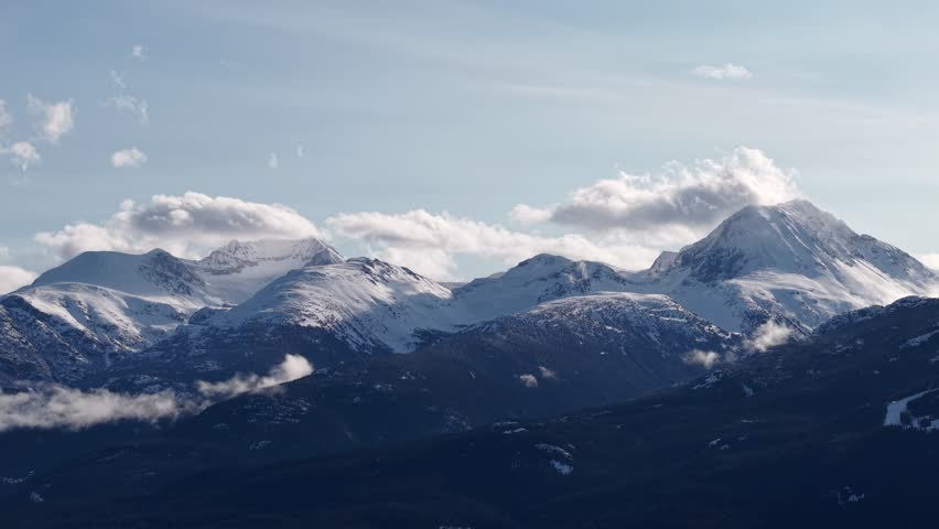 A breathtaking panoramic view of the Canadian wilderness featuring snow-covered mountains, dense evergreen forests, and a calm glacial lake. Captured under natural daylight with dramatic clouds, this 