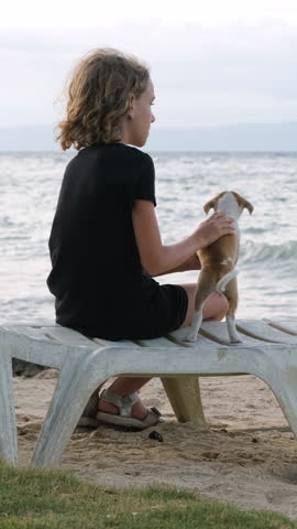 Vertical video. A young girl relaxes on a sunbed and pets an adorable puppy at sunset, gentle ocean waves lap against the shore, adding to the serene atmosphere