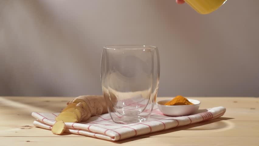 Golden turmeric latte pours into glass on cozy kitchen table with fresh ginger and turmeric powder, creating warm morning mood.