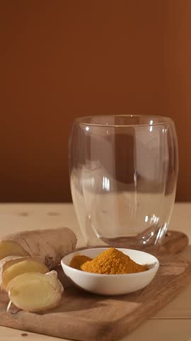 Golden turmeric latte poured into clear glass, ginger root and turmeric powder on wooden board, warm brown background adds cozy mood.