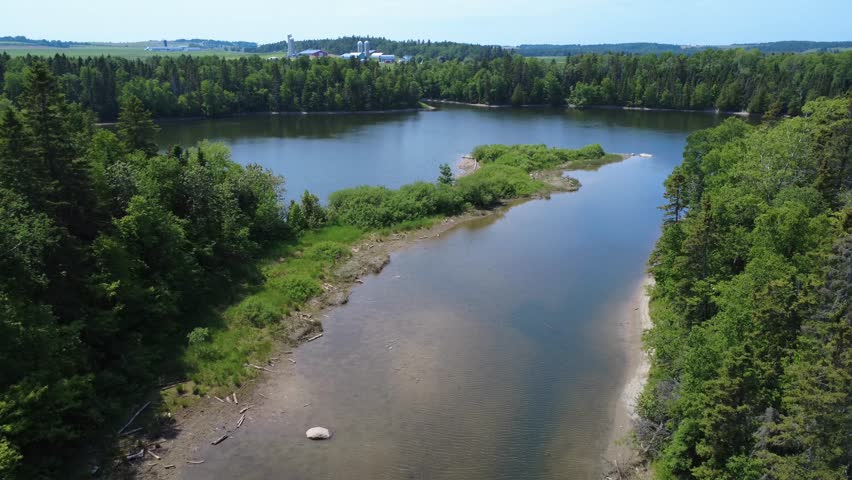 Aerial view of a calm river bordered by lush forest with a farm and hydroelectric dam in the background. Mitis River, Mitis-2 Generating Station, Quebec, Canada, 2025.