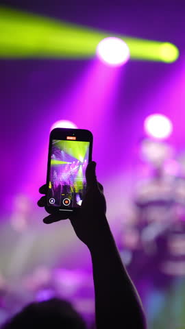A hand is holding a smartphone, capturing a lively concert scene filled with colorful lights and energetic performers in the background