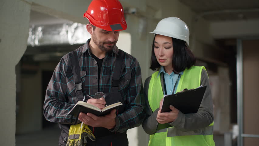 Multinational serious engineers discussing construction solutions project decisions planning review Asian woman pointing Caucasian man writing notebook teamwork infrastructure strategy renovation idea