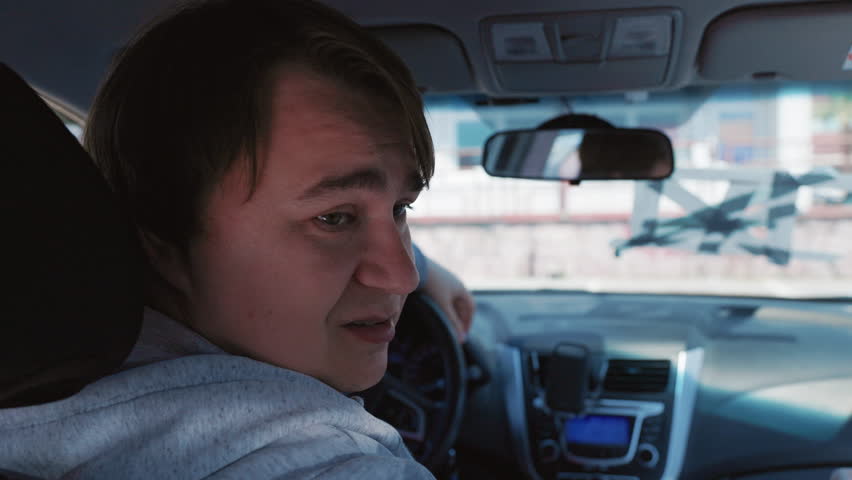 A man with short brown hair sitting in the driver seat of a car looks over his shoulder toward the back seat, with a steering wheel and dashboard partially visible