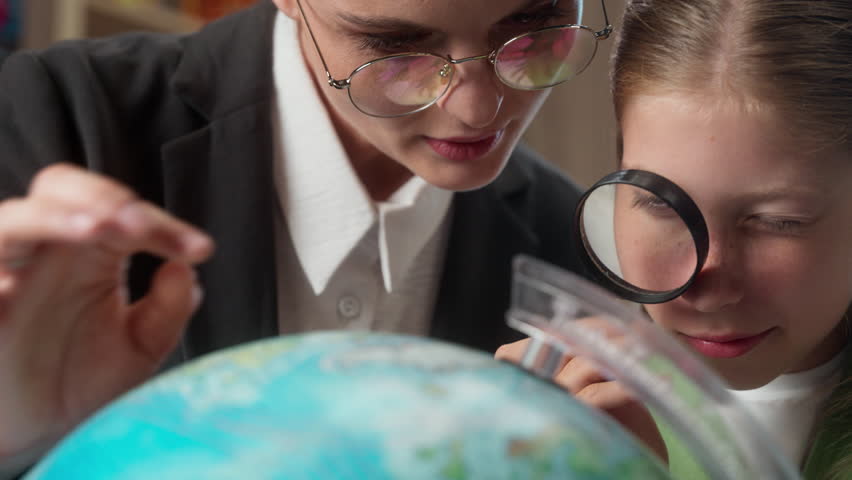 Caucasian teacher female woman professor explaining geography to little girl student school child kid schoolgirl looking through magnifying glass globe earth lens inspecting world map education indoor