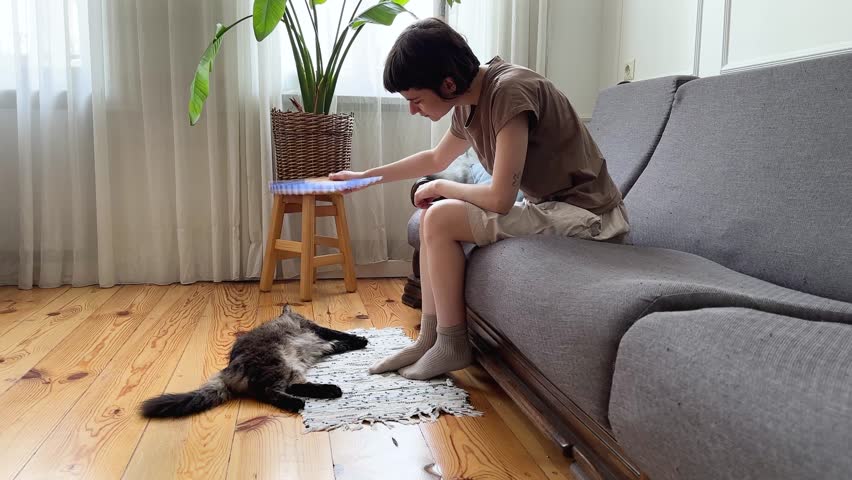 Young woman fanning cat to provide relief at home without AC. Pet care under high temperatures, shared discomfort, indoor climate struggle, animal wellbeing, cooling efforts. Heatwave, summer weather.