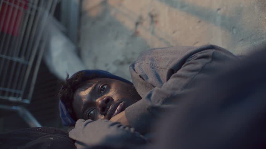 Young Black homeless man lying on city street, resting his head on arm and shivering from cold. Close-up view
