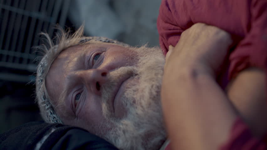 Elderly homeless man lying on sidewalk with eyes open and arms folded, staring pensively at people on street. Close-up shot