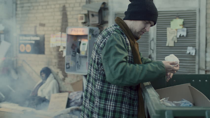 Homeless man digging through trash bin on urban street, beggar wrapped in blanket sleeping near payphone and people passing by in background
