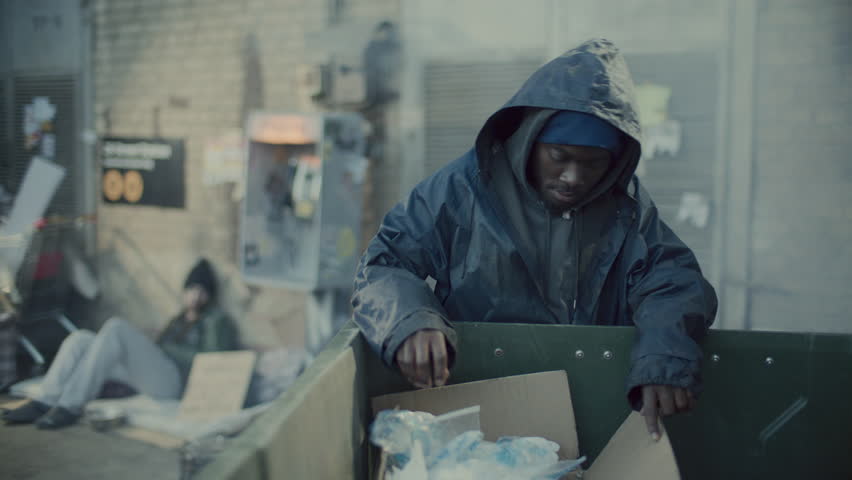 Homeless Black man in hooded jacket digging through dumpster in urban alleyway as people walking by in background