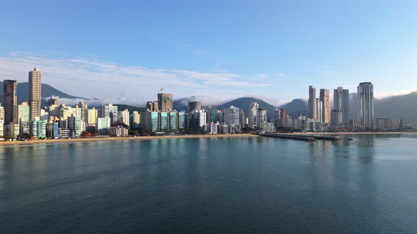 Panoramic drone movement of modern coastal Itapema settlement with high-rise buildings and mountain ranges behind the Meia Praia beach, Paraná, Brazil.