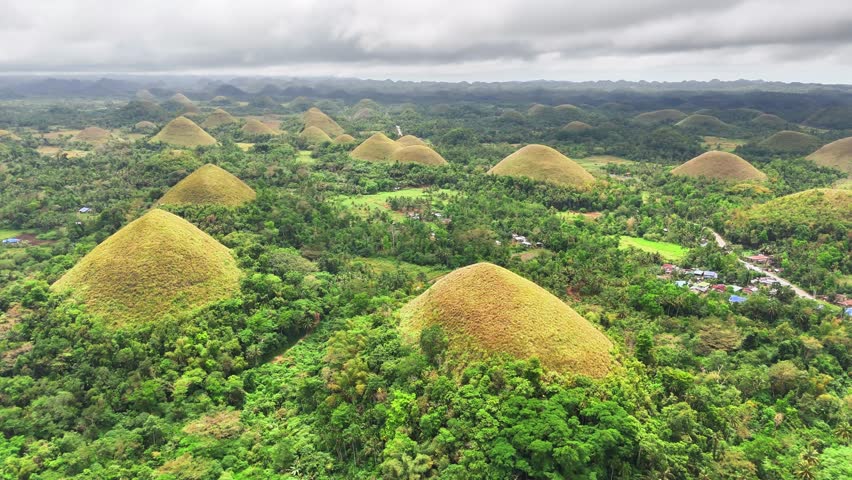 Aerial view of the Chocolate Hills in Bohol, Philippines, featuring conical grass-covered mounds rising from dense green forest, with hundreds of hills stretching across the horizon under cloudy skies