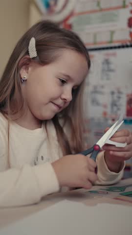 A young girl sits at a table and carefully cuts paper with scissors, focused on her creative project.