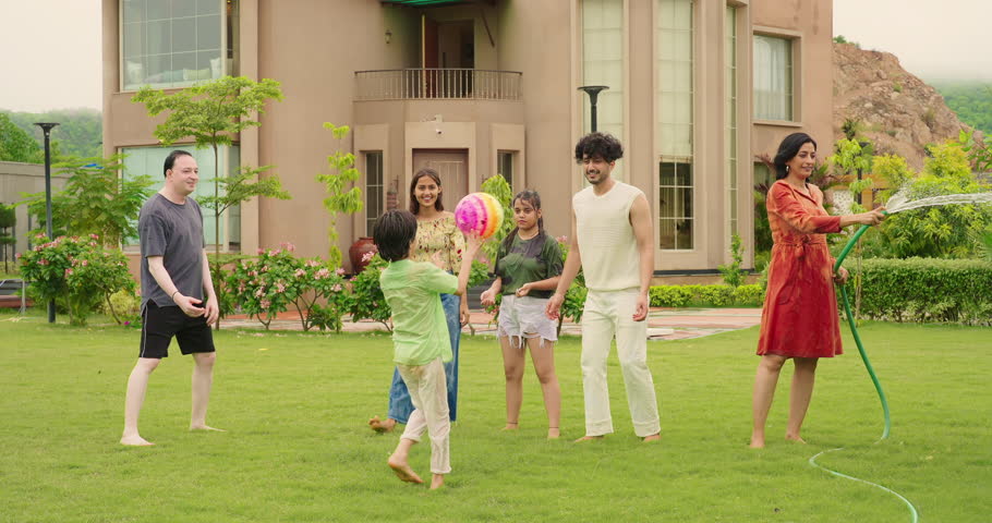 Indian happy mother hold hose pipe spraying water on extended family group playing games in outdoor house backyard lawn have fun joy enjoy day spend funny time summer vacation together outside park 