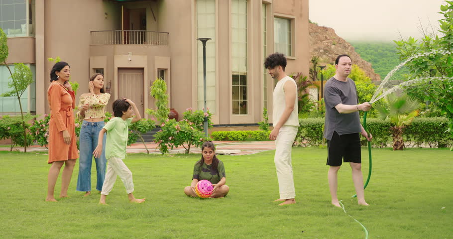 Indian happy father hold hose pipe spraying water on extended family group playing games in outdoor house backyard lawn having fun joy spend funny time enjoying summer vacation together outside park