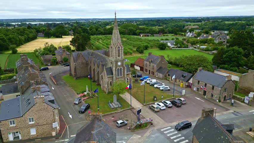 Drone advances in slight downward angle toward Notre-Dame-du-Guildo church and square in Saint-Cast-le-Guildo, with flags and surrounding fields. brittany in France