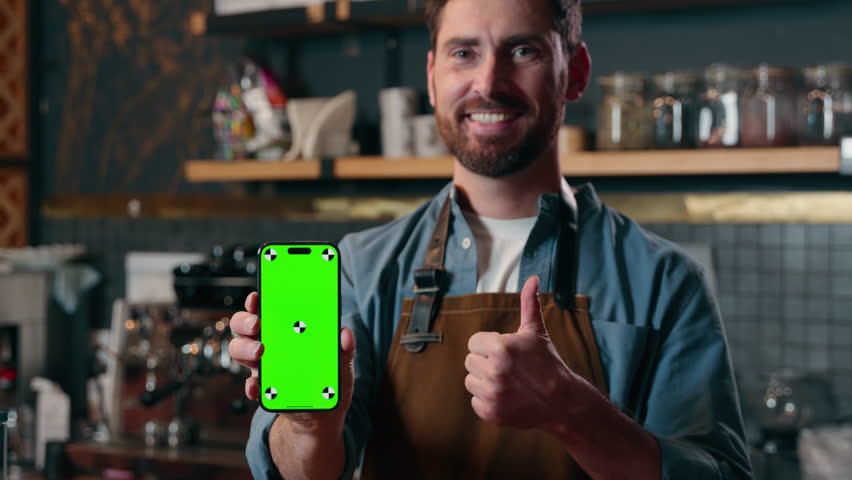 Satisfied happy Caucasian barista man showing green screen mobile phone thumb up in cafeteria cafe male small business entrepreneur recommend application advertisement guy waiter approval catering app