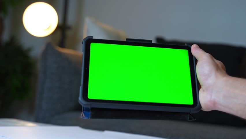 Hand holds tablet with green screen in warm home setting. Ideal for app previews, digital mockups, content creators, and tech promo scenes.