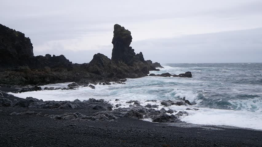 Dark waves crash against rocks on a black pebble beach in Iceland. The moody atmosphere and dramatic coastline reflect the raw power of nature.