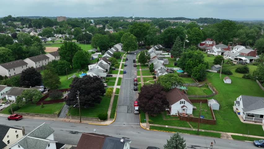 Quiet American suburb with identical single-family homes, tree-lined streets, green lawns and peaceful small-town atmosphere. Aerial flyover shot. Cloudy day in summer.