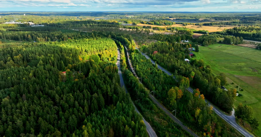 Aerial view following a long cargo train, in middle of sunny, lush green forest