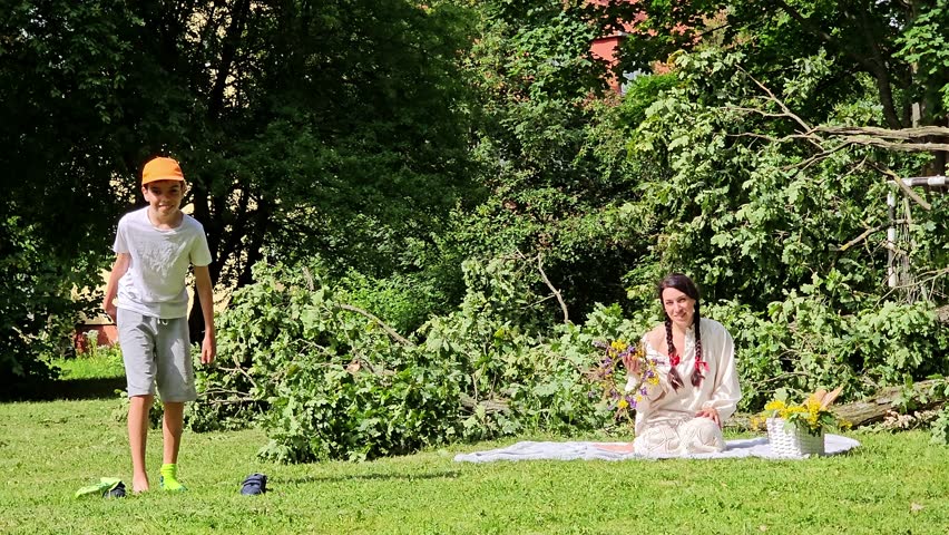 woman with son in white on grass picking berries, girl and boy in wreath with basket, outdoor recreation, travel, trip, mother and son picnic on grass, boy in orange cap takes off shoes