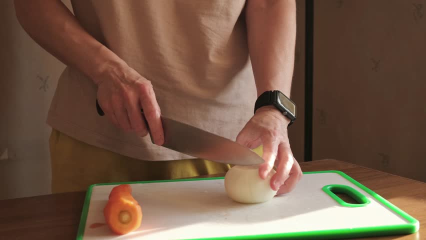 A person slicing a white onion on a plastic cutting board with a kitchen knife during food preparation at home, with soft natural light highlighting the hands and vegetables