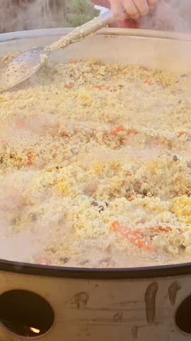 A flavorful rice dish simmers in a big pot at a lively community event as smoke rises into the evening air.