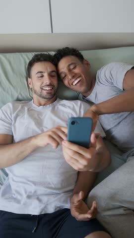 Gay couple sharing intimate moment, resting together, browsing smartphone with affection in cozy bedroom, enjoying peaceful morning connection